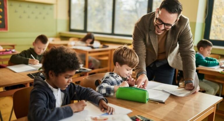 A group of students in the classroom. A teacher leans over a desk to help.