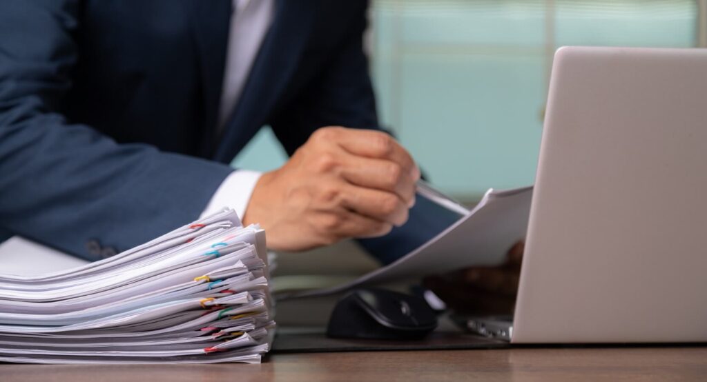 A close up of a pair of hands, holding a pen, with a computer on the desk